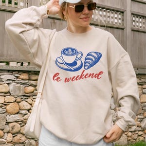 A woman wearing a white sweatshirt with a graphic design of a cup of coffee and a croissant, along with the text "Le weekend" printed on it.