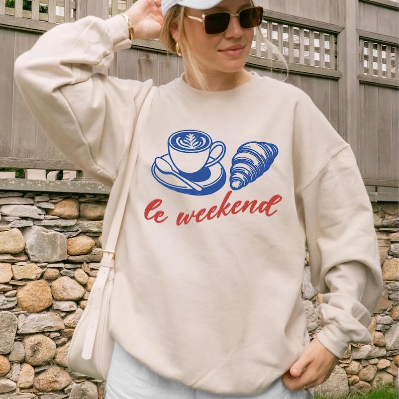 A woman wearing a white sweatshirt with a graphic design of a cup of coffee and a croissant, along with the text "Le weekend" printed on it.