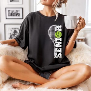 A woman sitting on a couch, wearing a black t-shirt with a tennis racket design and holding a white mug.