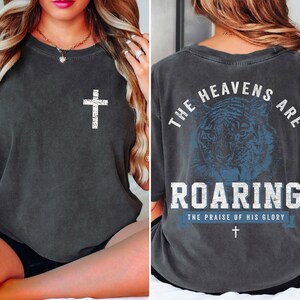 The image is a collage of two photos. In the left photo, a woman is sitting on a bed with her hair in a ponytail. In the right photo, the back of a gray t-shirt is shown, featuring a graphic design of a tiger and the text "The heavens are roaring" along with the phrase "The praise of his glory".