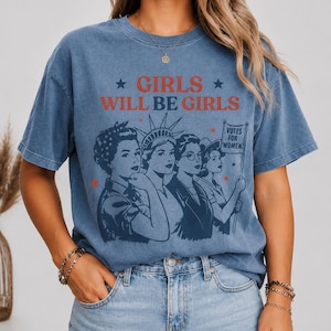 A woman wearing a blue t-shirt with a graphic design featuring a group of women and the text "GIRLS WILL BE GIRLS" printed on it.