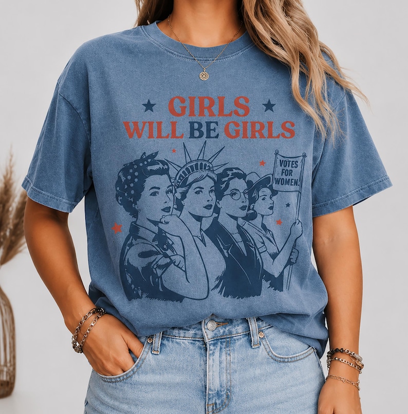 A woman wearing a blue t-shirt with a graphic design featuring a group of women and the text "GIRLS WILL BE GIRLS" printed on it.