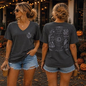 May include: A dark gray V-neck t-shirt featuring a small white graphic of a bottle on the front left chest. The back displays a large white print with celestial symbols, botanical illustrations, and text including "Always throw salt over your left shoulder."