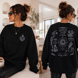 May include: A black long-sleeve sweatshirt featuring a small white graphic on the front chest and a large white mystical-themed design on the back. The back print includes a central sun and moon icon, botanical illustrations, and the text "Always throw spilled salt over your left shoulder," "Keep rosemary by your garden gate," "Plant roses and lavender for luck," "Fall in love whenever you can," "Live by nature," and "Stay wild."
