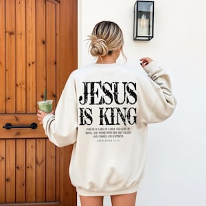 A woman wearing a white sweatshirt with the text "JESUS IS KING" printed on the back, standing in front of a wooden door.