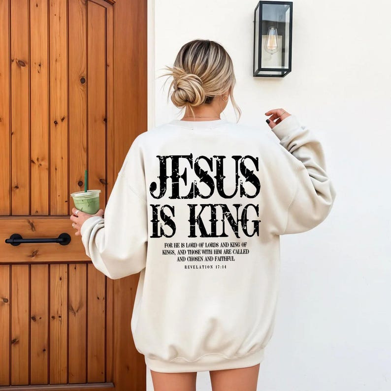 A woman wearing a white sweatshirt with the text "JESUS IS KING" printed on the back, standing in front of a wooden door.