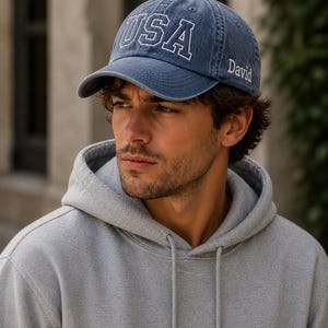 A man wearing a blue cap with the word "USA" printed on it, and a gray hoodie, looking to the side with a serious expression.