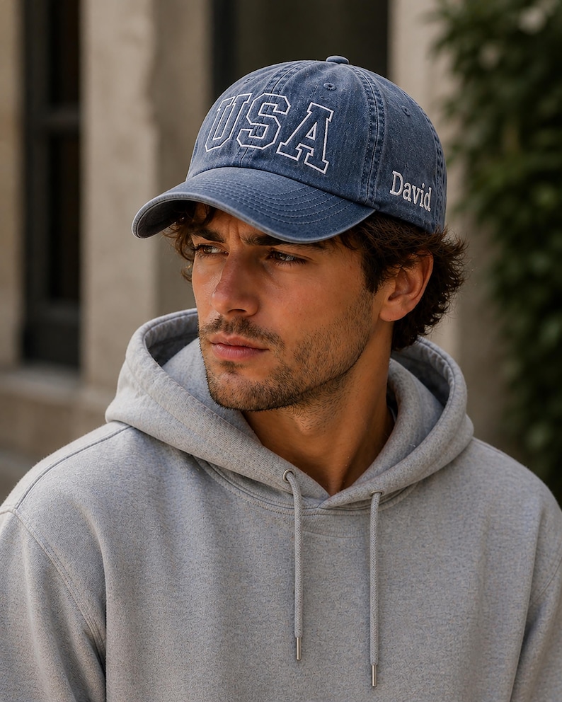 A man wearing a blue cap with the word "USA" printed on it, and a gray hoodie, looking to the side with a serious expression.