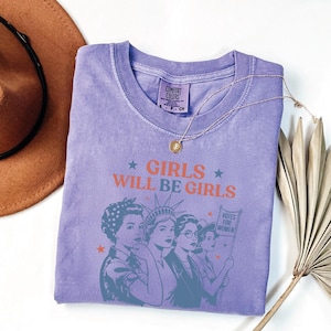 a purple t-shirt with a graphic design of three women, along with the text "GIRLS WILL BE GIRLS" printed on it. The t-shirt is laid out on a white surface, accompanied by a brown hat and a plant.