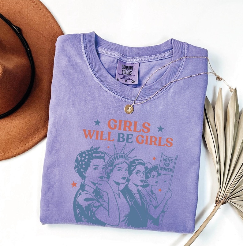 a purple t-shirt with a graphic design of three women, along with the text "GIRLS WILL BE GIRLS" printed on it. The t-shirt is laid out on a white surface, accompanied by a brown hat and a plant.