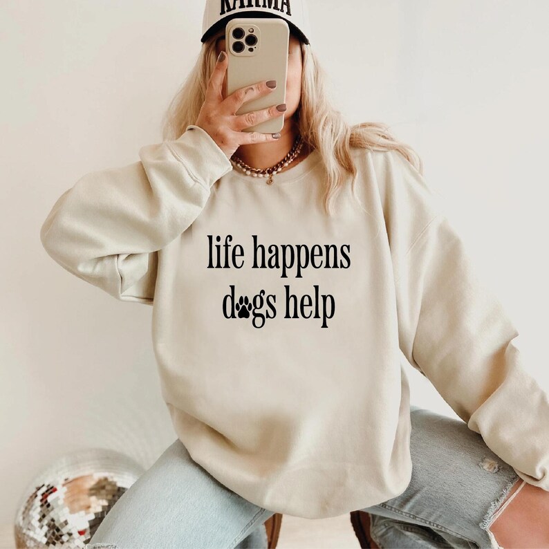 A person is sitting down and taking a selfie with a smartphone, wearing a sweatshirt with the text "life happens dogs help" printed on it.