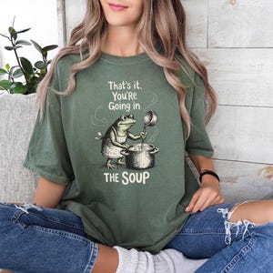 A woman wearing a green t-shirt with a cartoon frog and the text "That's it, You're going in the soup" is sitting on a couch.