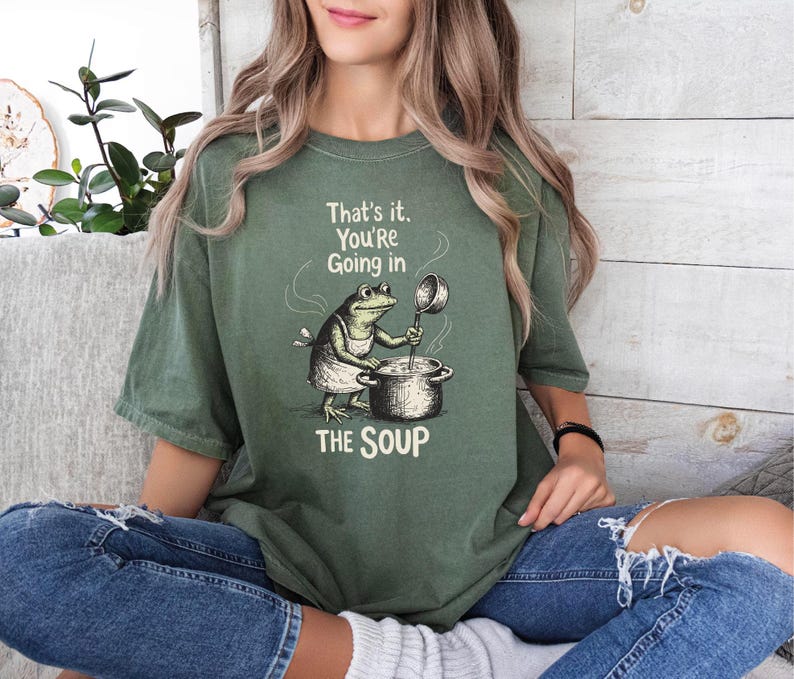 A woman wearing a green t-shirt with a cartoon frog and the text "That's it, You're going in the soup" is sitting on a couch.