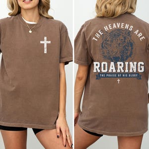 a woman wearing a brown t-shirt with a graphic design on the back. The design features a cross and the text "The heavens are roaring" along with the phrase "The praise of his glory".