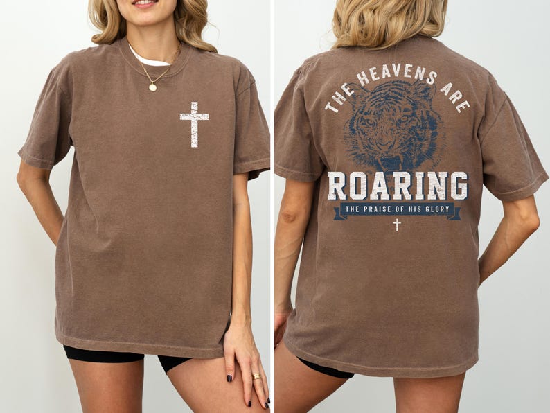 a woman wearing a brown t-shirt with a graphic design on the back. The design features a cross and the text "The heavens are roaring" along with the phrase "The praise of his glory".