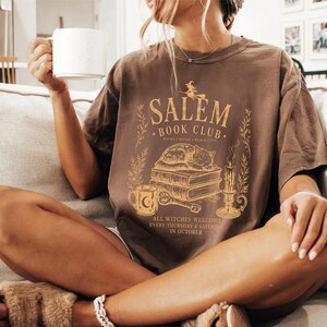 A woman sitting on a couch, holding a coffee mug and wearing a t-shirt with a graphic design.