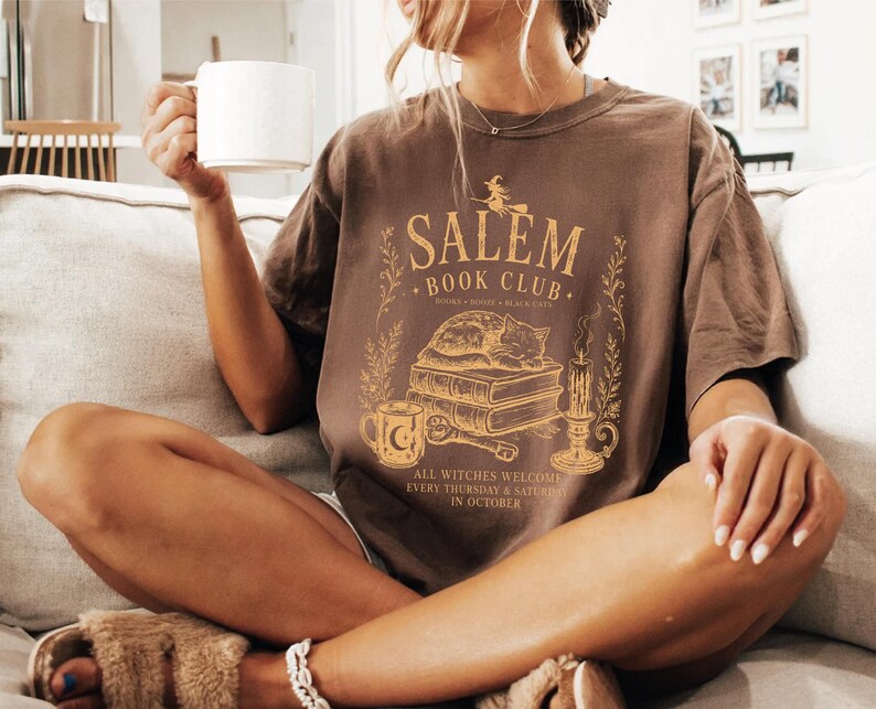 A woman sitting on a couch, holding a coffee mug and wearing a t-shirt with a graphic design.