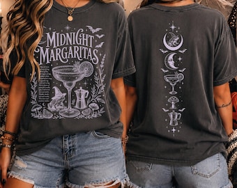 May include: A charcoal gray short-sleeve t-shirt featuring a white graphic print. The front displays the text "MIDNIGHT MARGARITAS" above a margarita glass, a frog wearing a witch hat, and a coffee pot. The back shows a vertical design of a moon, a margarita glass, a mushroom, and a coffee pot.