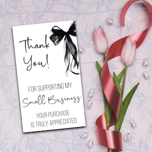 a white card with a black ribbon and pink tulips, along with a red ribbon and small white flowers, all arranged on a light pink background.