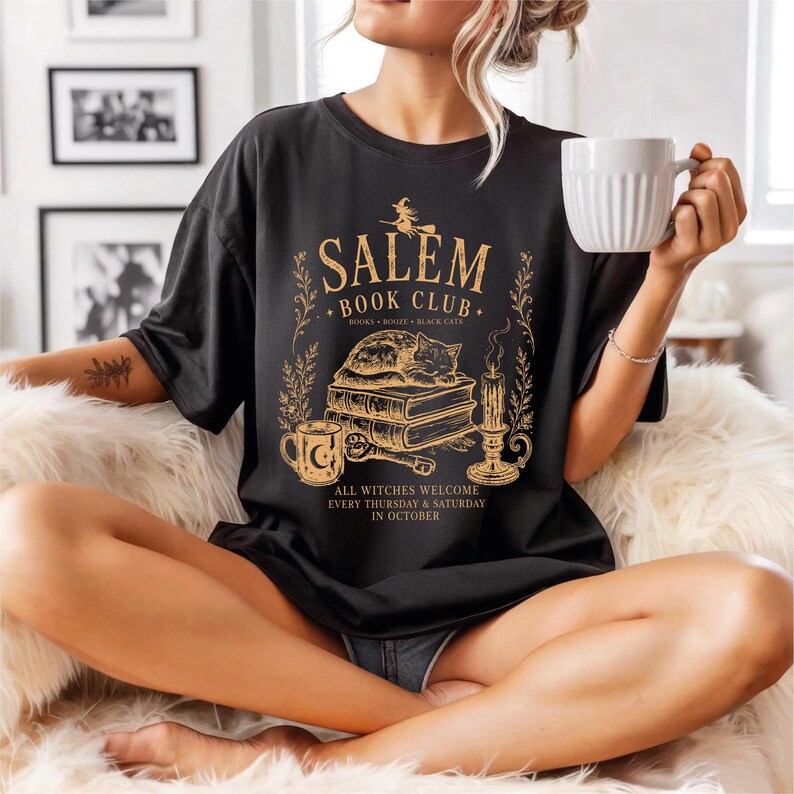A woman wearing a black t-shirt with a graphic design and text, sitting on a couch and holding a white mug.