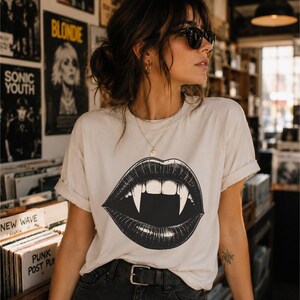 May include: A cream-colored short-sleeve t-shirt featuring a large black and white graphic print of open lips with prominent vampire fangs. The shirt has a crew neckline and rolled sleeves, paired with a black belt worn over dark-colored pants.