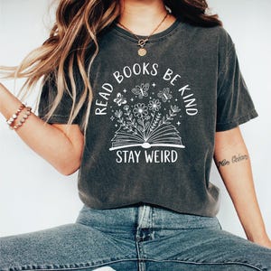 A woman wearing a gray t-shirt with a graphic design and text printed on it, sitting cross-legged with her hair blowing in the wind.
