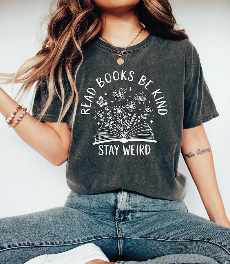 A woman wearing a gray t-shirt with a graphic design and text printed on it, sitting cross-legged with her hair blowing in the wind.