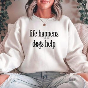 A woman wearing a white sweatshirt with the text "life happens dogs help" printed on it.