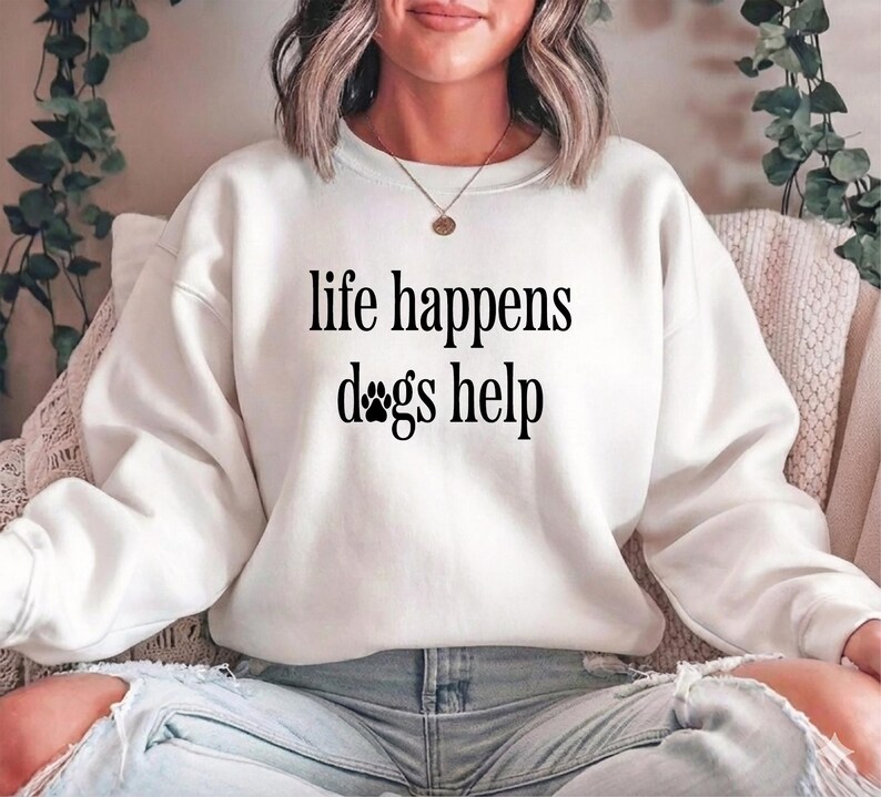 A woman wearing a white sweatshirt with the text "life happens dogs help" printed on it.