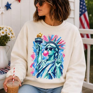 May include: A cream-colored crewneck sweatshirt featuring a vibrant graphic print of the Statue of Liberty wearing sunglasses and blowing a pink bubblegum bubble, holding a torch in one hand and making a peace sign with the other hand.