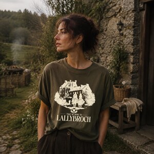 A woman wearing a t-shirt with a graphic design stands in front of a stone building, with a lush, forested background.