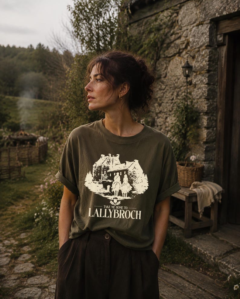 A woman wearing a t-shirt with a graphic design stands in front of a stone building, with a lush, forested background.