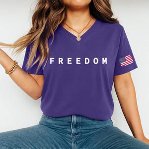 A woman wearing a purple t-shirt with the word "FREEDOM" printed on it, along with an American flag patch on the sleeve.