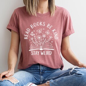 A woman wearing a pink t-shirt with the text "READ BOOKS BE KIND STAY WEIRD" printed on it, sitting cross-legged on the floor.