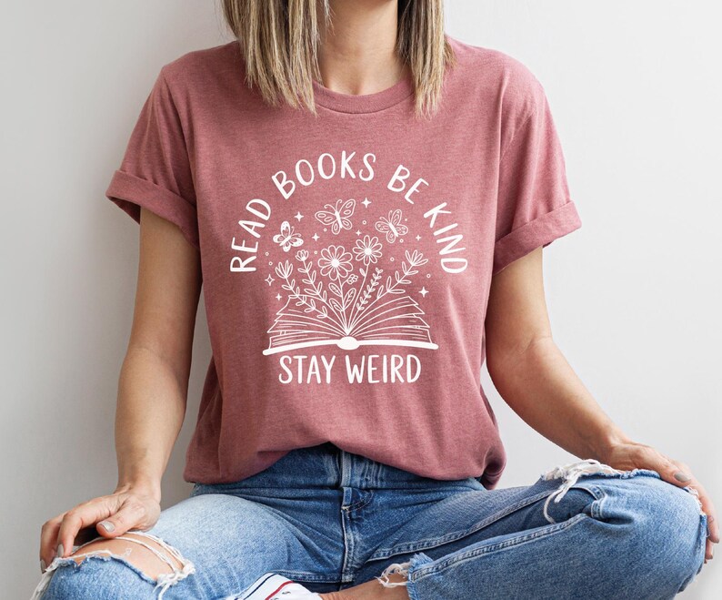 A woman wearing a pink t-shirt with the text "READ BOOKS BE KIND STAY WEIRD" printed on it, sitting cross-legged on the floor.