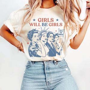 A woman wearing a white t-shirt with a graphic design featuring four women and the text "GIRLS WILL BE GIRLS" printed on it.