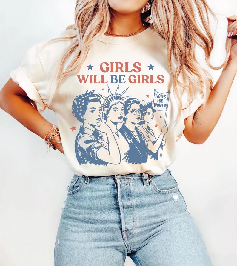 A woman wearing a white t-shirt with a graphic design featuring four women and the text "GIRLS WILL BE GIRLS" printed on it.