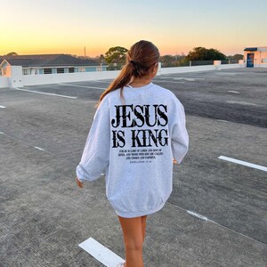 A person wearing a grey sweatshirt with the text "JESUS IS KING" printed on it, walking on a concrete surface with a sunset in the background.