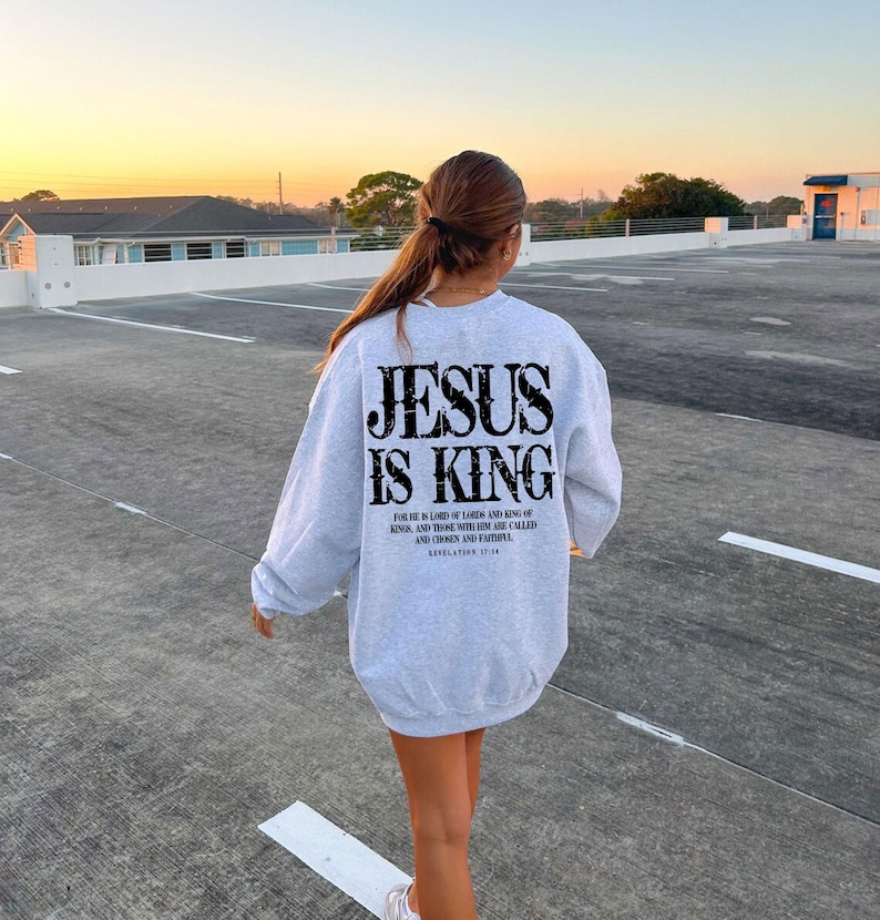 A person wearing a grey sweatshirt with the text "JESUS IS KING" printed on it, walking on a concrete surface with a sunset in the background.