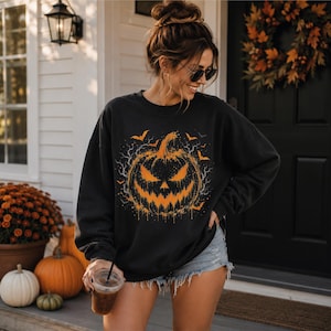 May include: A black long-sleeve crewneck sweatshirt featuring a large, central graphic of a distressed, orange jack-o'-lantern with a jagged, dripping smile, surrounded by small orange bats and thin, spindly gray branches on a dark background.