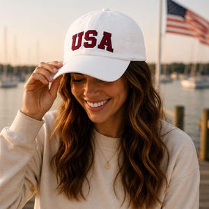 A woman wearing a white baseball cap with the word "USA" on it, smiling and looking down at her hat.
