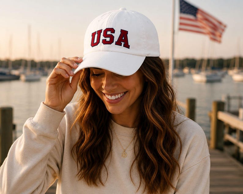 A woman wearing a white baseball cap with the word "USA" on it, smiling and looking down at her hat.