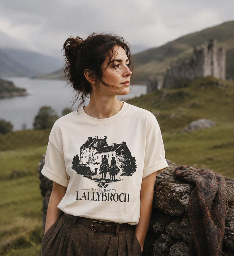 May include: Cream-colored t-shirt featuring a black graphic of a castle with two riders on horseback and the text "TAKE ME HOME TO LALLYBROCH." The shirt is paired with brown trousers and a brown belt. The background shows a scenic landscape.