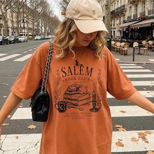 A woman wearing a cap and an orange t-shirt with a graphic design stands on a city street, with trees and buildings visible in the background.