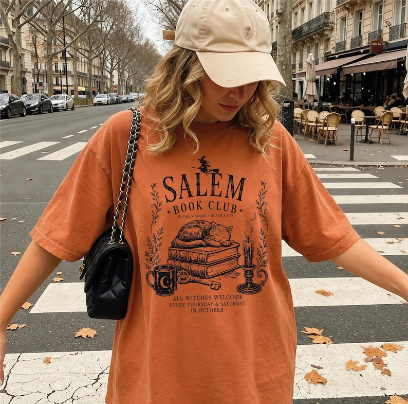 A woman wearing a cap and an orange t-shirt with a graphic design stands on a city street, with trees and buildings visible in the background.