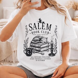 A woman wearing a white t-shirt with a graphic design featuring a witch, books, and a cat, sitting on a bed in a cozy bedroom setting.
