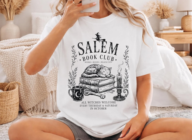 A woman wearing a white t-shirt with a graphic design featuring a witch, books, and a cat, sitting on a bed in a cozy bedroom setting.
