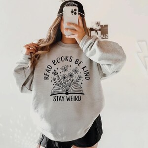 A person is taking a selfie with a white phone, wearing a gray sweatshirt with a graphic design and text printed on it.