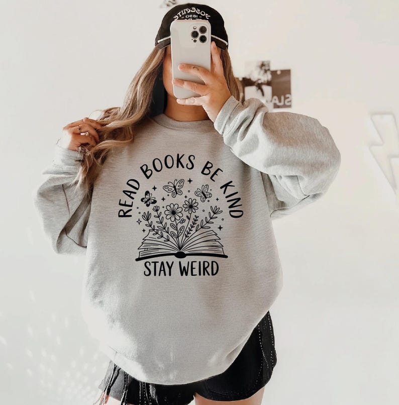 A person is taking a selfie with a white phone, wearing a gray sweatshirt with a graphic design and text printed on it.