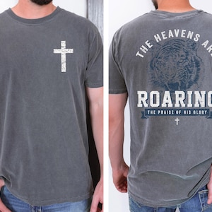 a man wearing a gray t-shirt with a graphic design on the back. The design features a cross and a tiger, along with the text "The heavens are roaring" and "The praise of his glory".